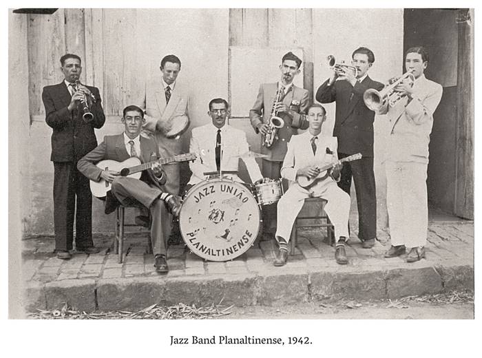  4 b, Jazz Band Planaltina 1942 Um grupo de homens todos vestidos de ternos posam para a foto, em que seguram equipamentos musicais, violão, viola, saxofone. Ao centro, um homem de terno está sentado, com as mãos sobre a bateria, onde aparece escrito: Jazz União Planaltinense. Realização: Academia Planaltinense de Letras, Artes e Ciências (APLAC), pelo Ecomuseu Pedra Fundamental e pelo Coletivo Nativo Audiodescrição produzida  pelo Instituto de Promoção das Pessoas com Deficiência Visual Audiodescritora: Elma Lúcia Rodrigues Consultor: Fernando Rodrigues Este projeto é promovido com recursos do Fundo de Apoio a Cultura do DF